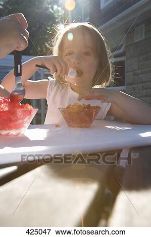 Stock Photo - Young girl eating jello at a table in a backyard. Fotosearch