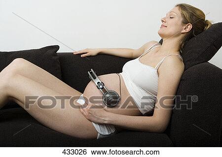 Stock Photograph - Pregnant woman lying down on a sofa with headphones on her stomach. Fotosearch