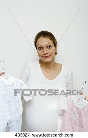Stock Photo - Pregnant woman shrugging whilst holding up blue and pink baby outfits. Fotosearch