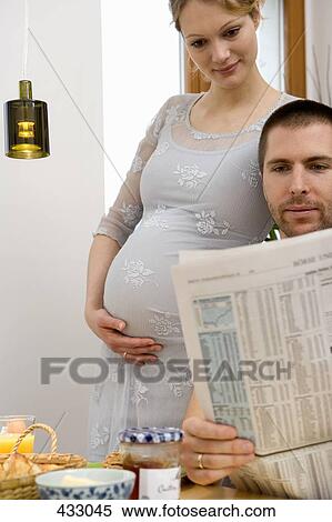 Stock Photography - Pregnant woman standing next to a man reading a newspaper over breakfast. Fotosearch