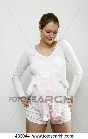 Picture - Pregnant woman with a pink baby outfit on her stomach. Fotosearch