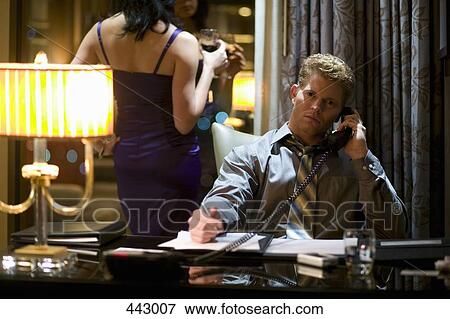 Stock Photo - Businessman sitting at a desk using a telephone whilst a woman stands behind holding a glass of wine. Fotosearch