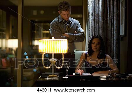 Businesswoman sitting at a desk and signing a document with a man standing behind her View Large Photo Image Stock Image - Businesswoman sitting at a desk and signing a document with a man standing behind her. Fotosearch