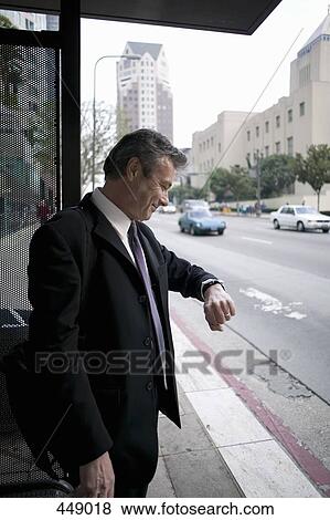 Stock Photo - Businessman looking at his watch whilst standing at a bus stop. Fotosearch