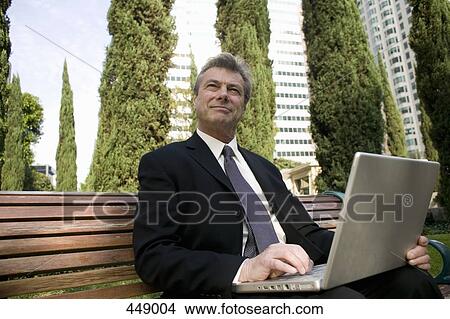 Picture - Businessman sitting on a park bench and using a laptop. Fotosearch