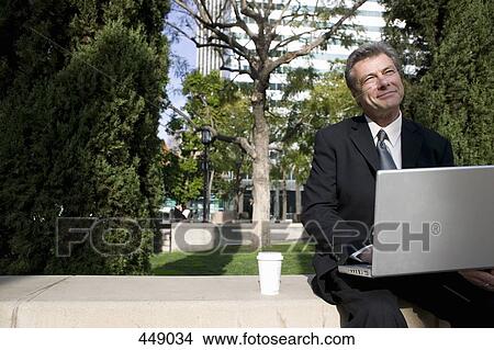 Businessman sitting on a park bench and using a laptop View Large Photo Image Picture - Businessman sitting on a park bench and using a laptop. Fotosearch