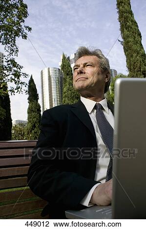 Stock Image - Businessman sitting on a park bench and using a laptop. Fotosearch