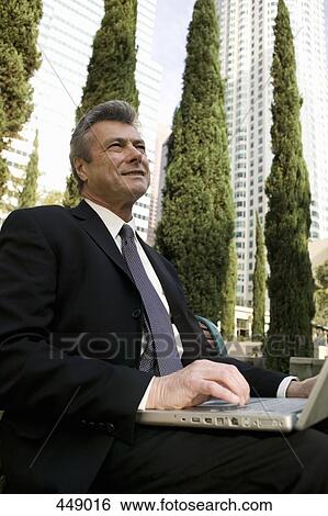 Businessman sitting on a park bench and using a laptop View Large Photo Image Stock Photograph - Businessman sitting on a park bench and using a laptop. Fotosearch