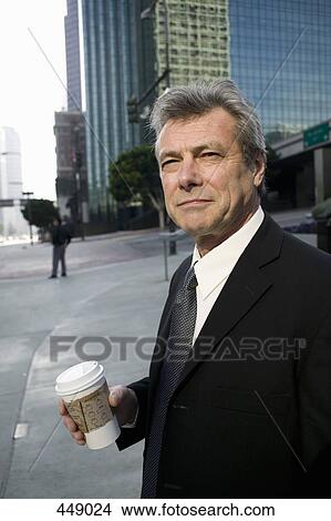 Businessman standing on a street corner holding a cup of coffee View Large Photo Image Picture - Businessman standing on a street corner holding a cup of coffee. Fotosearch