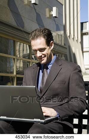 Businessman sitting on an outside bench using a laptop View Large Photo Image Picture - Businessman sitting on an outside bench using a laptop. Fotosearch