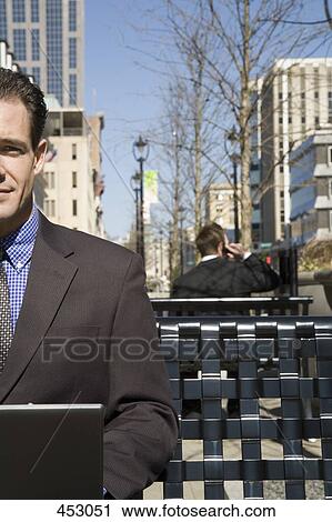 Businessman sitting on an outside bench using a laptop View Large Photo Image Stock Image - Businessman sitting on an outside bench using a laptop. Fotosearch