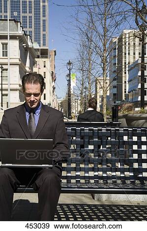 Businessman sitting on an outside bench using a laptop View Large Photo Image Stock Photo - Businessman sitting on an outside bench using a laptop. Fotosearch