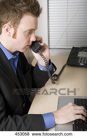 Young businessman on the phone and using a laptop whilst sitting at a desk View Large Photo Image Stock Photo - Young businessman on the phone and using a laptop whilst sitting at a desk. Fotosearch