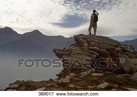 Stock Photo - Man standing on a rocky ledge and looking at the mountains. Fotosearch