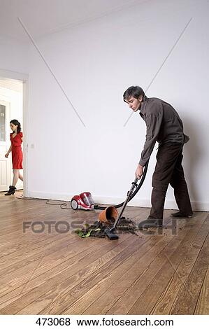 Stock Photo - A man vacuuming up soil from a spilt potted plant whilst a woman is walking down a hallway. Fotosearch
