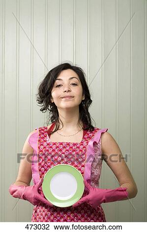 Stock Image - A woman holding a plate whilst wearing an apron and rubber gloves. Fotosearch