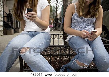 Stock Image - Two adolescent girls sitting on railing and using mobile phones. Fotosearch