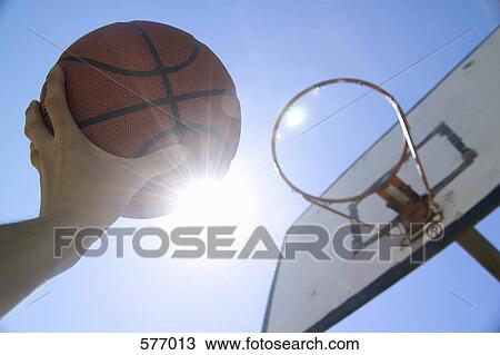 Stock Image - A human hand reaching up towards the sun and basketball hoop with holding basketball. Fotosearch