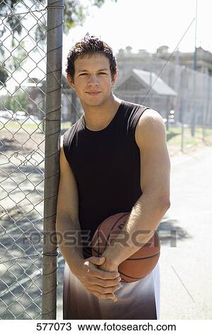 Stock Image - A young man leaning against a chainlink fence with a basketball. Fotosearch