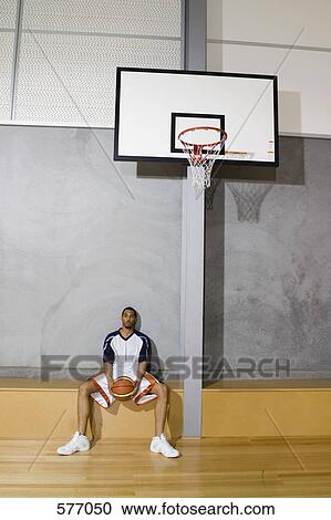 Stock Image - A young man sitting and holding a basketball between his legs. Fotosearch