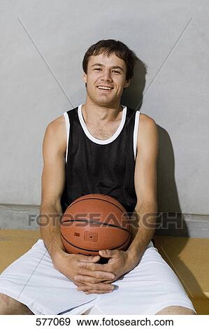 Stock Photo - A young man sitting on a bench with arms around a basketball. Fotosearch