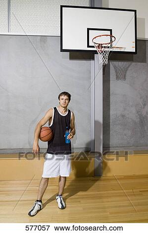 Stock Photo - A young man standing on a basketball court holding a sports drink and a basketball. Fotosearch