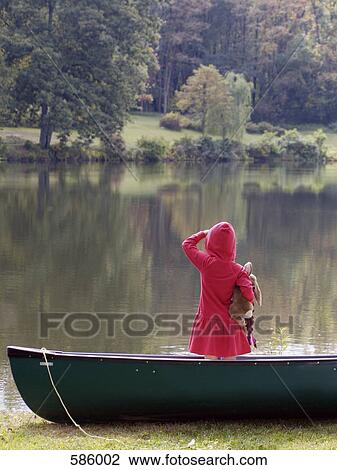 Stock Image - Rear View of a child holding a stuffed toy standing on a beached boat looking across a lake. Fotosearch