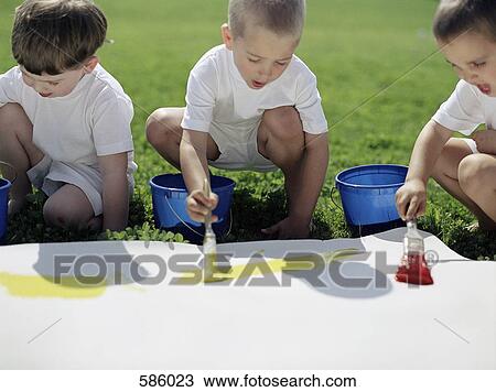 Stock Image - Three boys painting on paper outside. Fotosearch