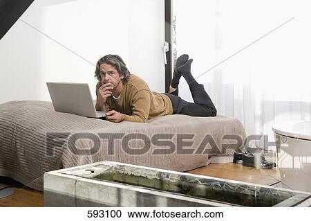 A man lying on a bed and using a laptop View Large Photo Image Stock Image - A man lying on a bed and using a laptop. Fotosearch