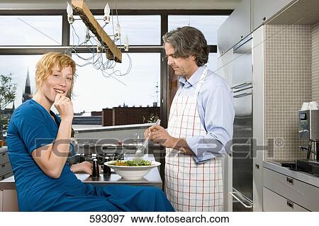 Stock Photo - A man preparing a meal in a kitchen with a woman sitting near. Fotosearch