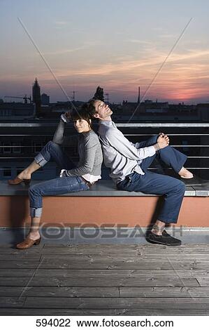 Stock Image - A couple sitting back to back on a rooftop terrace at night. Fotosearch