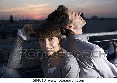 Stock Photograph - A couple sitting back to back on a rooftop terrace at night. Fotosearch