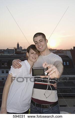 Stock Image - A young couple photographing themselves on a rooftop terrace. Fotosearch