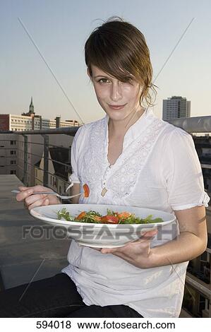 A young woman sitting on a rooftop terrace holding a plate of salad View Large Photo Image Stock Photo - A young woman sitting on a rooftop terrace holding a plate of salad. Fotosearch