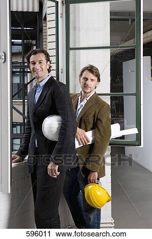 Two men standing by a window holding hardhats and architectural plans View Large Photo Image Stock Image - Two men standing by a window holding hardhats and architectural plans. Fotosearch
