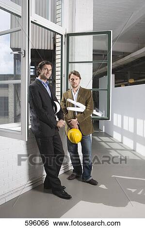 Two men standing by a window holding hardhats and architectural plans View Large Photo Image Stock Photograph - Two men standing by a window holding hardhats and architectural plans. Fotosearch