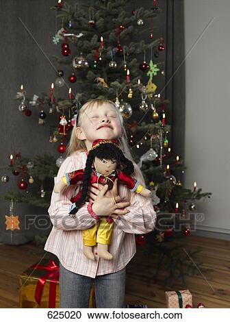 Stock Image - A girl embracing a doll in front of a Christmas tree. Fotosearch