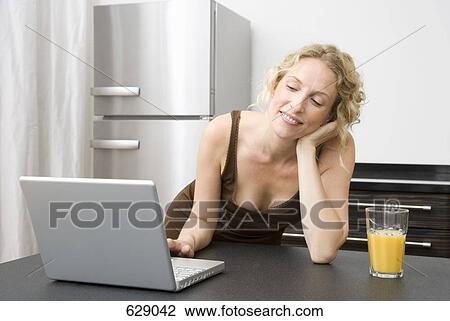 Stock Image - A mid adult woman leaning on a kitchen counter using a laptop. Fotosearch