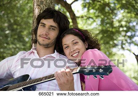 Stock Image - A young couple sitting underneath a tree and playing a guitar. Fotosearch