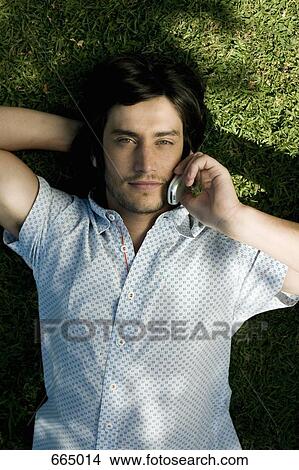 Picture - A young man lying on grass and using his mobile phone. Fotosearch
