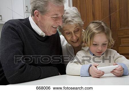 A grandson using a electronic device while his grandparents watch View Large Photo Image Stock Photograph - A grandson using a electronic device while his grandparents watch. Fotosearch