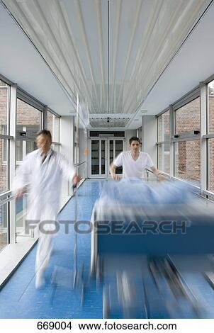 Picture - Two healthcare workers pushing a hospital bed along a corridor. Fotosearch