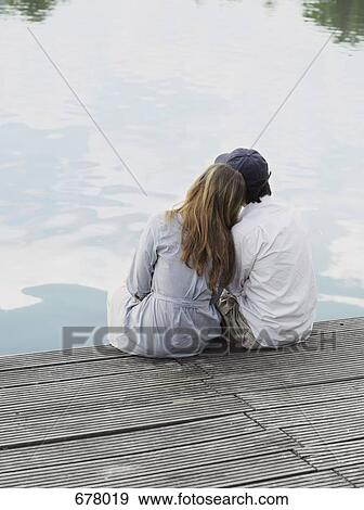 Stock Photo - A young couple sitting on a jetty, rear view. Fotosearch