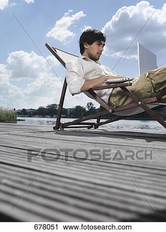 Stock Image - A young man sitting in a deck chair on a jetty using a laptop. Fotosearch