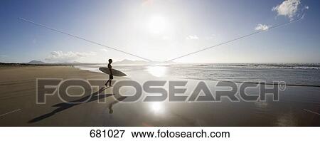 A young man standing on a beach and carrying a surfboard View Large Photo Image Stock Photo - A young man standing on a beach and carrying a surfboard. Fotosearch