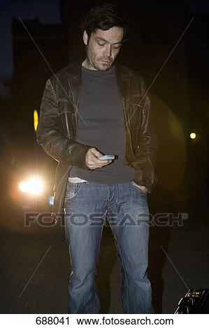 Stock Image - A man standing in front of headlights looking at his mobile phone. Fotosearch