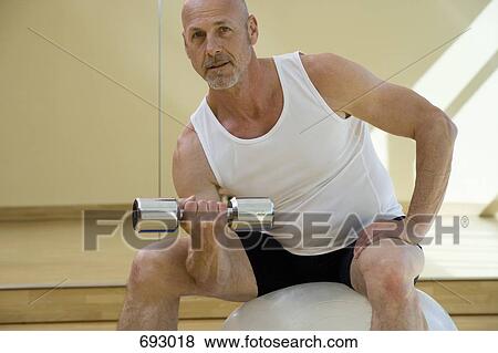 A man sitting on an exercise ball lifting a dumbbell View Large Photo Image Stock Photo - A man sitting on an exercise ball lifting a dumbbell. Fotosearch