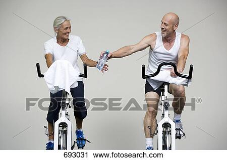 Stock Image - A senior woman and a mature man riding stationary bikes and passing a water bottle. Fotosearch