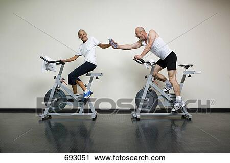 A senior woman and a mature man riding stationary bikes and passing a water bottle View Large Photo Image Stock Image - A senior woman and a mature man riding stationary bikes and passing a water bottle. Fotosearch