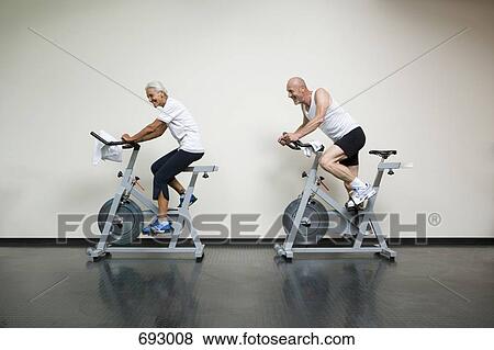 Stock Photo - A senior woman and a mature man riding stationary bikes. Fotosearch
